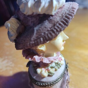May include: A decorative figurine of a woman wearing a large hat and a pink dress. The hat is brown with a white trim. The figurine has a detailed face and is set on a light-colored base. The overall color scheme is soft and pastel.