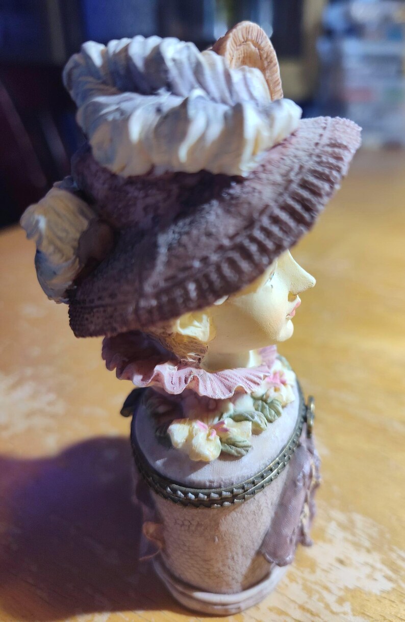 May include: A decorative figurine of a woman wearing a large hat and a pink dress. The hat is brown with a white trim. The figurine has a detailed face and is set on a light-colored base. The overall color scheme is soft and pastel.