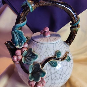 May include: A white ceramic teapot with a crackled glaze and a vine design. The teapot has a brown handle and a lid with pink flowers.