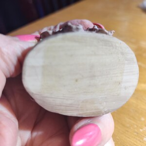May include: A light-colored, wooden, oval-shaped object with a textured surface. The top edge features a pink and brown decorative element. The object is held in a hand with pink nail polish.