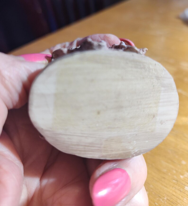 May include: A light-colored, wooden, oval-shaped object with a textured surface. The top edge features a pink and brown decorative element. The object is held in a hand with pink nail polish.