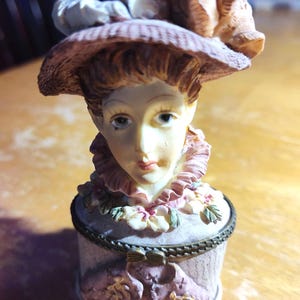 May include: A decorative figurine of a woman wearing a hat, with a detailed face and a ruffled collar. The figurine is painted in soft pastel colors, with a small, round container base. The hat and container are adorned with floral accents.