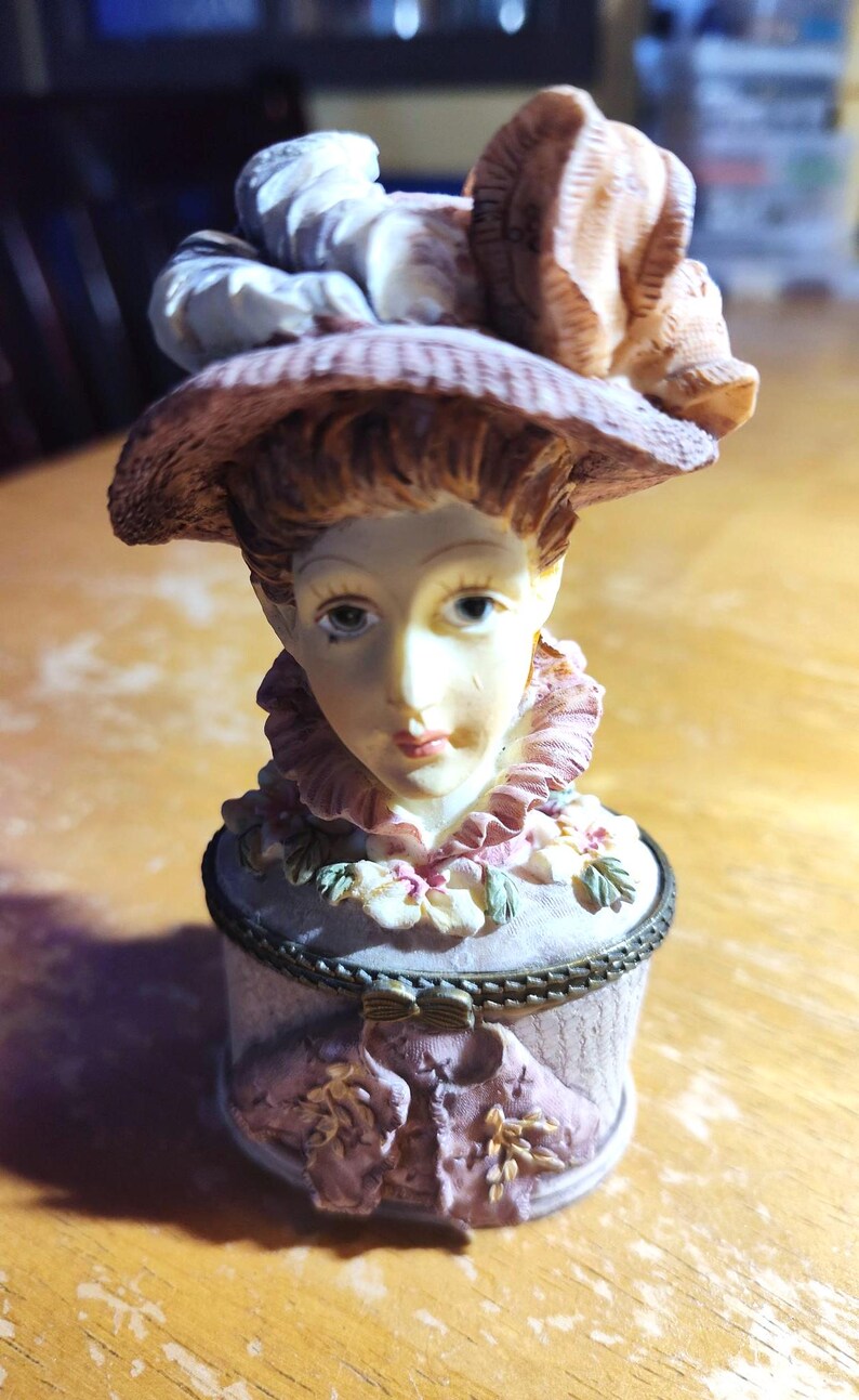 May include: A decorative figurine of a woman wearing a hat, with a detailed face and a ruffled collar. The figurine is painted in soft pastel colors, with a small, round container base. The hat and container are adorned with floral accents.