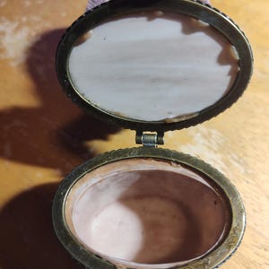 May include: An open, antique oval trinket box with a bronze-colored metal frame. The interior is a pale pink, and the lid is lined with a matching fabric. The box is hinged and has a decorative clasp. The box is likely made of porcelain or ceramic.