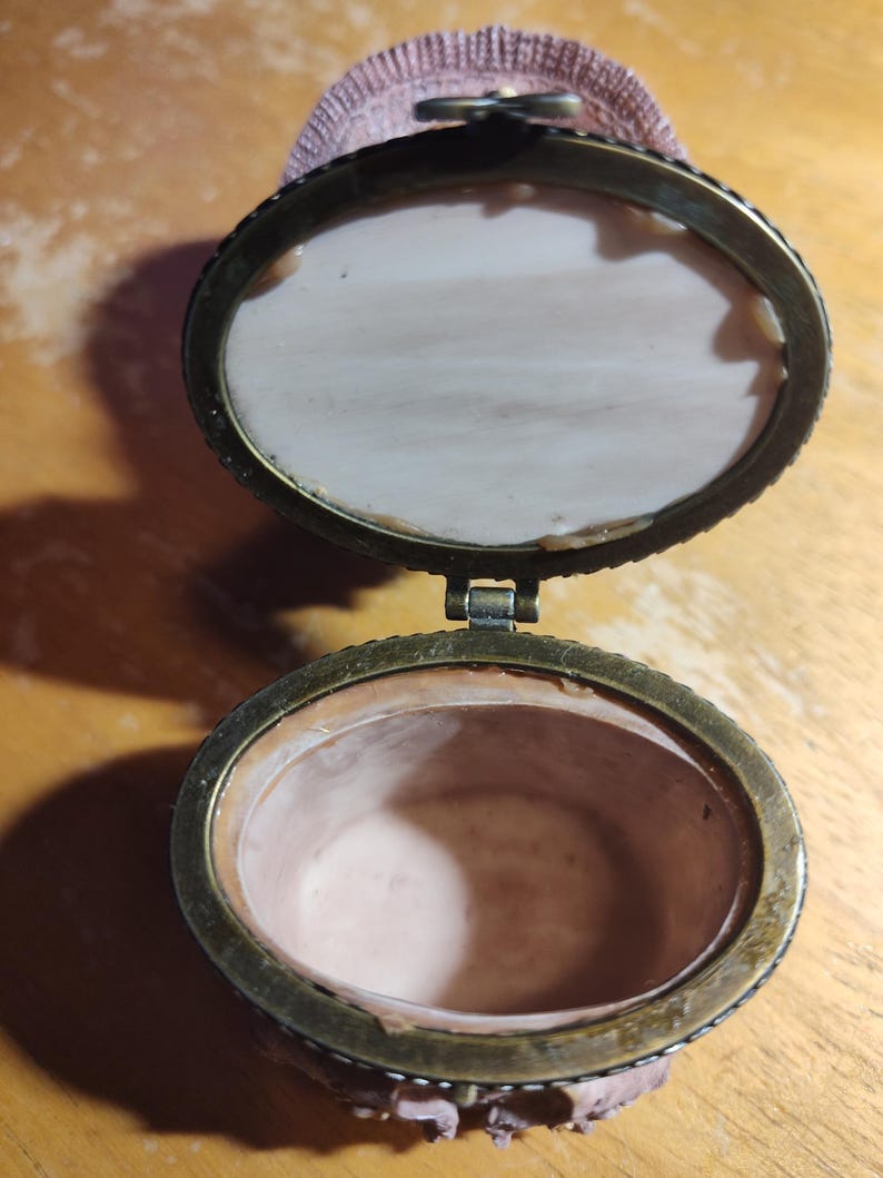 May include: An open, antique oval trinket box with a bronze-colored metal frame. The interior is a pale pink, and the lid is lined with a matching fabric. The box is hinged and has a decorative clasp. The box is likely made of porcelain or ceramic.