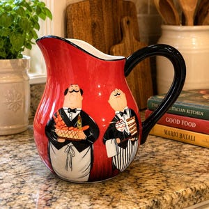 May include: A red ceramic pitcher featuring a black handle and a painted illustration of two waiters in black suits and white aprons. One waiter holds a tray of carrots and a bottle, while the other holds a tray with a dessert and a glass.