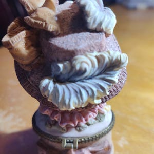 May include: A decorative trinket box with a hat-shaped lid. The hat is a mix of beige, pink, and white, with intricate details. The box itself is a light pink with floral accents and a gold-colored rim. A charming collectible.