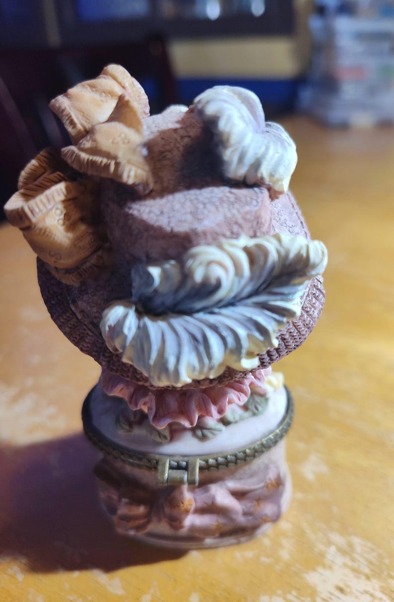 May include: A decorative trinket box with a hat-shaped lid. The hat is a mix of beige, pink, and white, with intricate details. The box itself is a light pink with floral accents and a gold-colored rim. A charming collectible.
