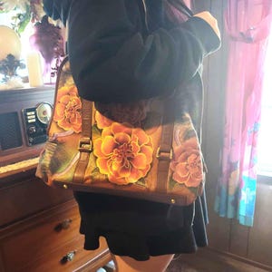 May include: A brown leather handbag with large, vibrant orange and pink floral designs. The bag has a rectangular shape with a top handle and a shoulder strap. It features a buckle closure and is carried over the shoulder.