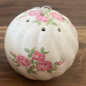May include: A spherical white ceramic pomander featuring a ribbed surface and a floral pattern of pink roses with green leaves. The top of the object has several small circular ventilation holes and a silver-colored metallic hanging cord.