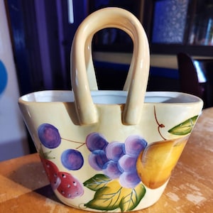May include: A ceramic basket-shaped container with a cream-colored exterior and a curved handle. The basket is decorated with painted purple grapes, a strawberry, and a yellow pear. The interior is white.