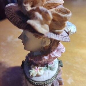 May include: A decorative figurine of a woman wearing a hat with floral accents. The figurine has a light skin tone, with a pink and white dress. The hat is brown and beige, and the figurine appears to be made of ceramic.