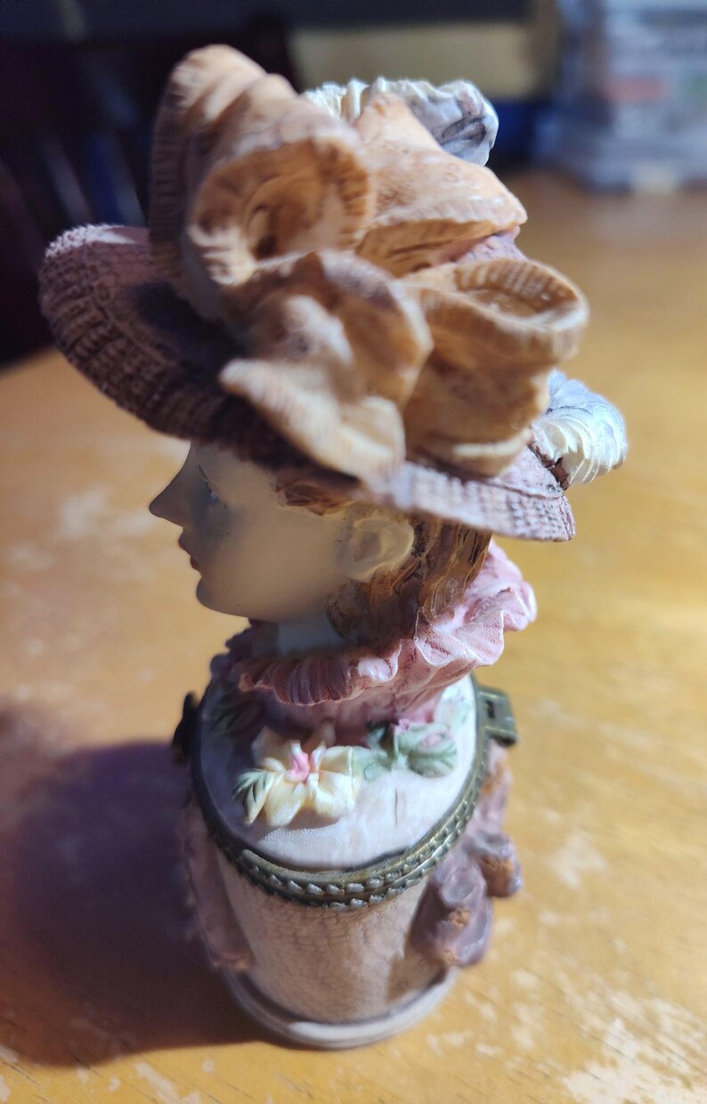 May include: A decorative figurine of a woman wearing a hat with floral accents. The figurine has a light skin tone, with a pink and white dress. The hat is brown and beige, and the figurine appears to be made of ceramic.