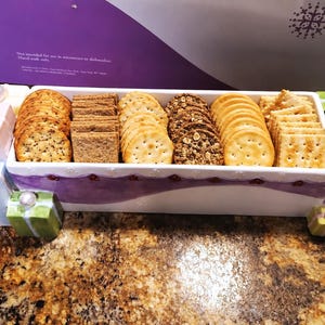 May include: A rectangular white ceramic serving tray filled with various crackers. The tray features a purple decorative band and is adorned with small, colorful gift-box-shaped accents at the corners, each topped with a white spherical bead.