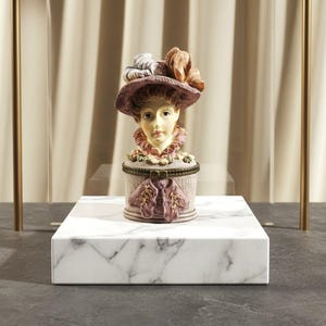 May include: A decorative trinket box featuring a woman's bust wearing a hat adorned with feathers. The box is light pink with a floral design and a bow clasp. The bust has a detailed face and is set on a white marble base.