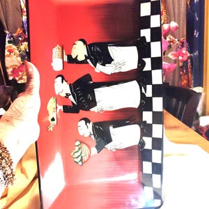 May include: Rectangular serving tray with a red background and a black and white checkered border. The tray features illustrations of three waiters in black suits and white aprons, each holding a different dish.