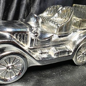 May include: A shiny silver-toned vintage automobile figurine featuring an open-top design with detailed bucket seats, a prominent front grille, spoked wheels, and a spare tire mounted on the rear. The metallic surface reflects light across its curved body.