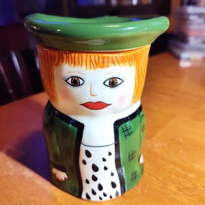 May include: A ceramic jar with a painted face, orange hair, and a green hat. The jar has a green jacket with black accents and a white shirt with black polka dots. The jar is sitting on a wooden surface.