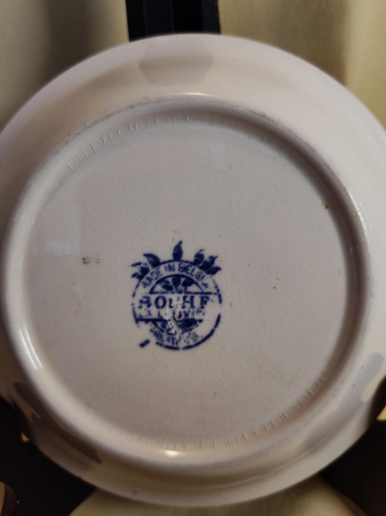 Peut inclure: Une assiette en c&eacute;ramique blanche avec un tampon bleu indiquant "MADE IN BELGIUM" et "SOUPLE". L'assiette a un bord l&eacute;g&egrave;rement sur&eacute;lev&eacute; et une surface lisse. Le tampon est au centre de l'assiette.