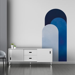 Puede incluir: Una cómoda blanca con dos cajones y una puerta corredera. La cómoda tiene un acabado de madera y está sentada en un suelo gris. La pared detrás de la cómoda es blanca y tiene un diseño de arco abstracto azul y gris.