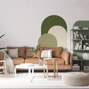May include: A living room interior with a brown leather sofa, two white coffee tables, a beige knitted pouf, and a green bookshelf with books and a fan. The wall behind the sofa features a green, beige, and cream abstract geometric design.