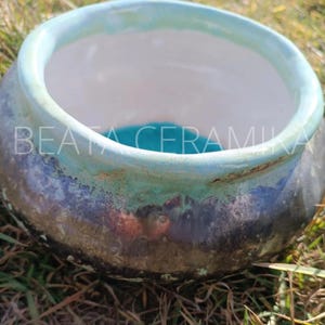 Może przedstawiać: Ceramiczna miska z turkusowym rantem i czarnym, zielonym i brązowym, nakrapianym szkliwem. Miska stoi na zielonej trawie.