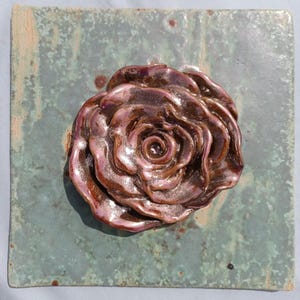Pode incluir: Um azulejo de parede de cerâmica quadrado com uma rosa esculpida em três dimensões num esmalte metálico brilhante em tons de bronze e roxo. O azulejo de fundo tem um acabamento manchado e desgastado em tons de verde sálvia e azul-petróleo.