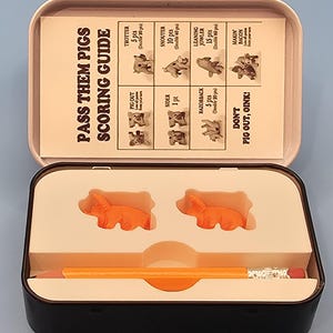 以下が含まれることがあります： 黒いブリキの箱に入ったPass Them Pigsゲーム。中には、2つのオレンジ色の豚のフィギュア、鉛筆、スコアリングガイドがあります。ガイドには豚の位置とポイント値が記載されています。「Pass Them Pigs Scoring Guide」の文字が見えます。