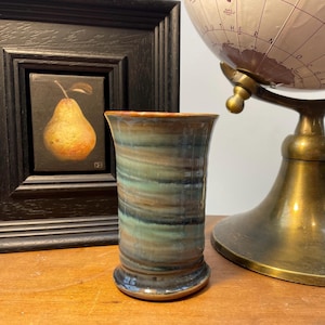May include: A handmade ceramic vase with a flared top and a circular base. The vase has a swirl pattern of green, brown, and blue tones. A framed painting of a pear and a brass globe are in the background.