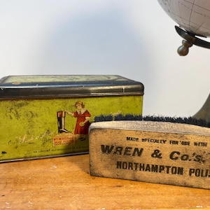 May include: A vintage green and yellow tin box with a picture of a girl polishing a boot. The box is labelled "Wren & Co.'s Northampton Polish". A wooden shoe brush with black bristles is in the foreground.