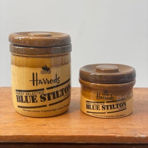 May include: Two brown and tan ceramic cheese jars with lids, labelled "Harrods Finest Selected Blue Stilton Cheese." The larger jar is taller than the smaller one. Both jars are on a wooden surface.