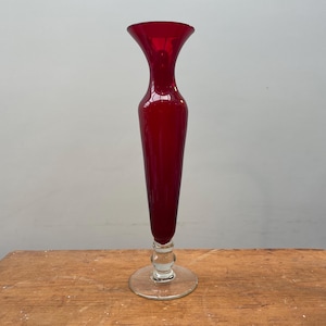 May include: A tall, ruby red glass vase with a flared top and a slender body. The vase has a clear base and a decorative stem. The vase is approximately 30 cm tall.