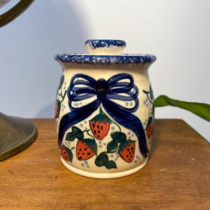 May include: A small ceramic jar with a lid, decorated with a blue bow and red strawberries on a white background.