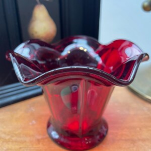 May include: A vibrant ruby red glass bowl with a wavy rim. The bowl features a pedestal base and a fluted design. The transparent glass allows light to pass through. Likely vintage, suitable for serving, and a decorative piece.