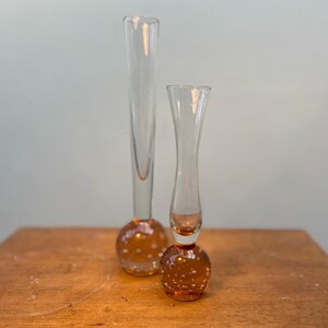 May include: Two clear glass bud vases. One is tall and slender, the other is shorter and wider. Both have a round, amber-coloured base with small air bubbles. The vases are on a wooden surface.
