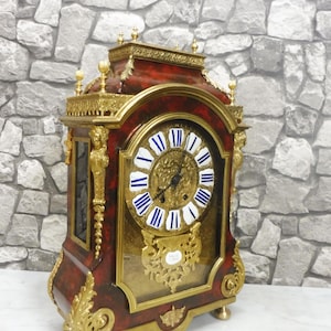 Op de afbeelding: Een schoorsteenklok met een donkerrode kunstmarmeren afwerking en sierlijke goudkleurige metalen accenten. De wijzerplaat heeft witte cirkelvormige markeringen met blauwe Romeinse cijfers, zwarte wijzers en een gouden decoratieve plaat.