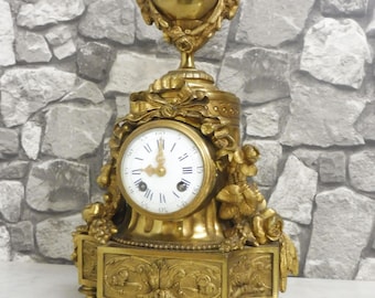 May include: Ornate gold-colored antique clock with a round clock face featuring Roman numerals. The clock has intricate floral and decorative details, including a vase-shaped top and a rectangular base with sculpted designs. The clock is a vintage decorative item.