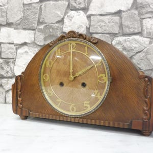 May include: A vintage wooden mantel clock with a light brown finish. The clock face is gold with black numerals and hands. The clock has a decorative design on the top and sides. The clock is approximately 12 inches wide and 8 inches tall.