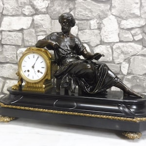 May include: An antique bronze mantel clock featuring a seated figure and a round clock face with Roman numerals. The clock has a black base with gold accents and decorative feet. The background is a stone wall.