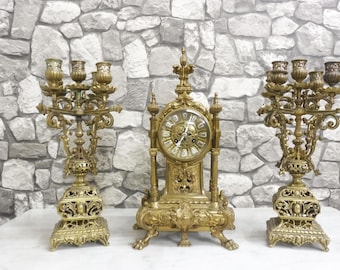 May include: An antique gold-colored clock flanked by two matching candelabras. The clock has a round face with Roman numerals and ornate detailing. The candelabras each have five candle holders and intricate designs, creating a classic, elegant aesthetic.