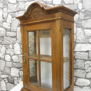 May include: A wooden wall-mounted display cabinet with glass doors and shelves. The cabinet has a carved design on the top and a brass lock on the door.