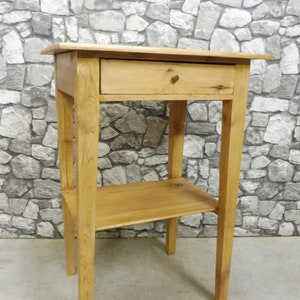 Op de afbeelding: Een licht houten bijzettafel met een enkele lade en een lager plank. De tafel heeft een eenvoudig, rustiek ontwerp en is gemaakt van natuurlijk hout.