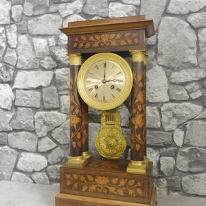 May include: An antique wooden clock with a gold-coloured face and pendulum. The clock features intricate floral inlay designs on the wooden frame and pillars. The clock's face has black Roman numerals and hands. The clock is a decorative item.