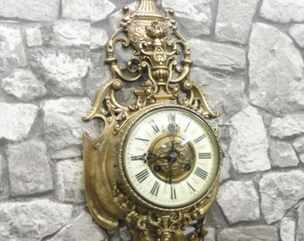 May include: Ornate antique wall clock with a gold-colored finish. The clock features a round face with Roman numerals and decorative scrollwork. The clock is mounted on a stone-textured wall.
