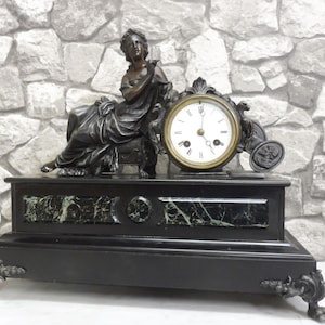 May include: Antique black mantel clock with a bronze-colored sculpture of a woman. The clock has a white face with black Roman numerals and ornate gold-colored hands. The base is black with green marble accents.