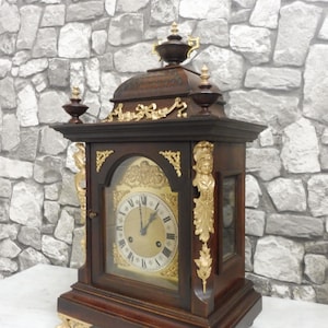 Könnte beinhalten: Eine Kaminuhr aus dunklem Holz mit goldenen Akzenten und einem Glaszifferblatt. Die Uhr hat ein dekoratives Oberteil und verzierte Füße.