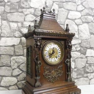 Puede incluir: Un reloj grande de madera adornado con una esfera dorada y números romanos. El reloj tiene un marco de madera tallada decorativo y un techo puntiagudo.