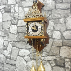 Puede incluir: Un reloj de pared de madera con esfera cuadrada, centro negro y números romanos blancos. El reloj está adornado con detalles metálicos dorados, una pequeña esfera azul en la parte superior y dos pesas colgantes doradas en forma de pera.