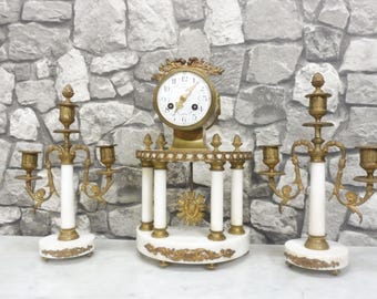 May include: A vintage clock and candelabra set. The clock has a gold-colored frame and a white face. The candelabras are white with gold-colored accents. The set is made of marble and metal. The clock is flanked by two candelabras.