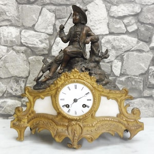 Op de afbeelding: Een schoorsteenklok met een bronskleurig figuur van een persoon die een lange staf vasthoudt, zittend op een decoratieve goudkleurige metalen voet. De wijzerplaat is wit met zwarte Romeinse cijfers en twee zwarte wijzers in een sierlijk frame.